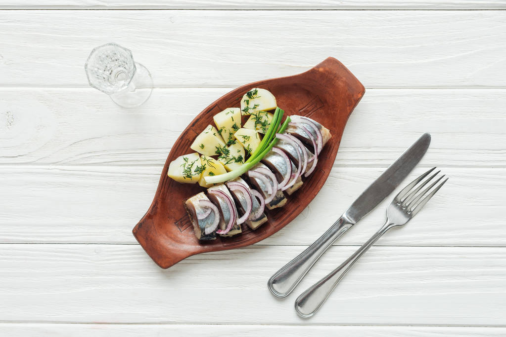 Widok z góry Śledź marynowany, ziemniaków i cebuli w talerz Ceramika z kieliszek wódki i sztućce na białym tle drewnianych - Zdjęcie, obraz
