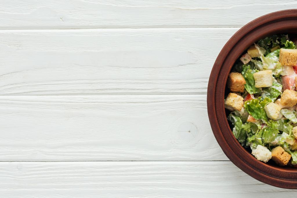 сверху вид на традиционный салат Цезарь с гренками в миске на белом деревянном фоне с копировальным пространством
 - Фото, изображение