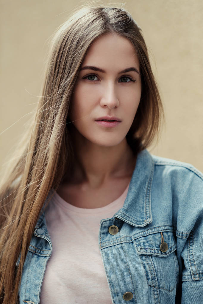 Young beautiful girl with long hair in a T-shirt and denim jacket on the background of vintage concrete wall. - Photo, Image
