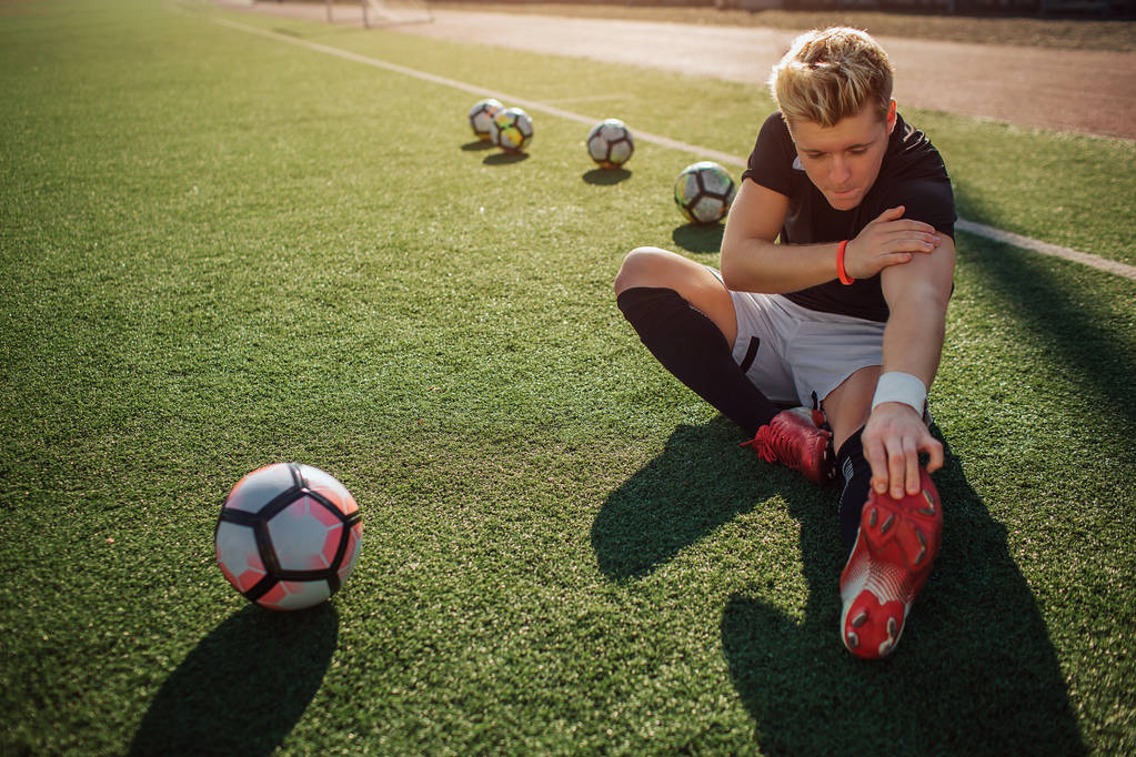 Mladá blondýnka fotbal hráče sit na trávníku a roztáhnout. Dosáhne jednou rukou, na nohou. Další je na rameni. Čtyři kuličky za ním a jeden míč v přední. - Fotografie, Obrázek