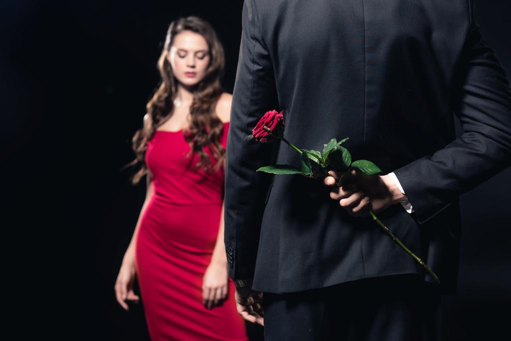 selective focus of man hiding rose behind back with woman in red dress on background isolated on black - Photo, Image