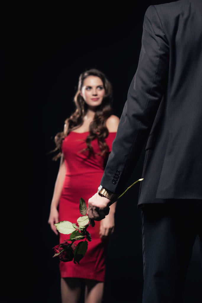 επιλεκτική εστίαση του ανθρώπου που κρατώντας τριαντάφυλλο με γυναίκας με κόκκινο φόρεμα σε απομονωμένα σε μαύρο φόντο - Φωτογραφία, εικόνα
