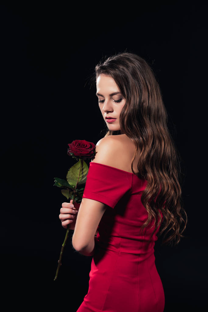 hermosa mujer en vestido rojo con los ojos cerrados sosteniendo rosa aislado en negro
 - Foto, imagen