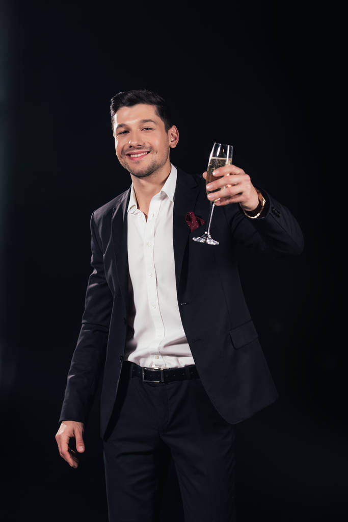 sorrindo bonito homem olhando para a câmera e levantando copo de champanhe isolado em preto
 - Foto, Imagem