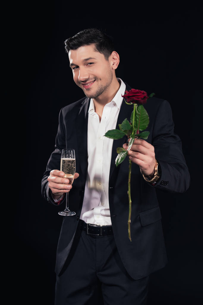 smiling handsome man looking at camera and holding glass of champagne with red rose isolated on black - Photo, Image