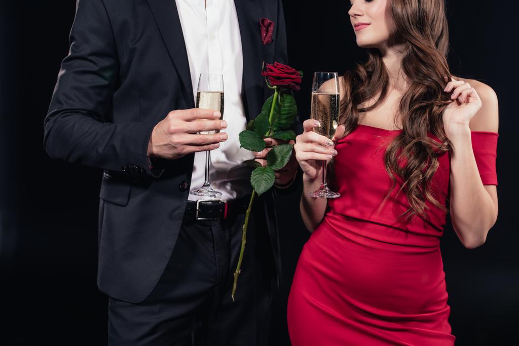 cropped view of couple holding champagne glasses and red rose isolated on black - Photo, Image