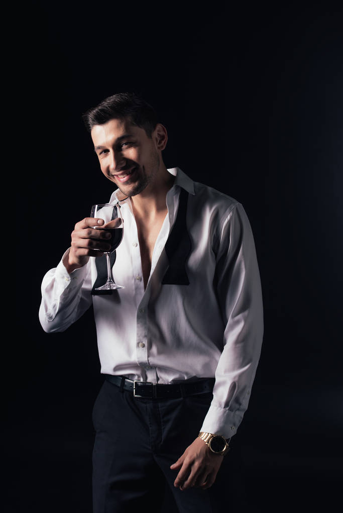 usmívající se muž v bílé košili, při pohledu na fotoaparát a držení sklenice červeného vína izolované na černém - Fotografie, Obrázek