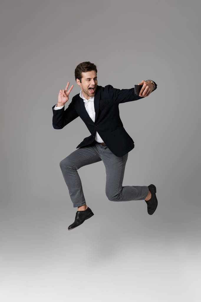 Retrato de comprimento total de um jovem empresário encantado vestido de terno isolado sobre fundo cinza, tirando uma selfie enquanto salta
 - Foto, Imagem
