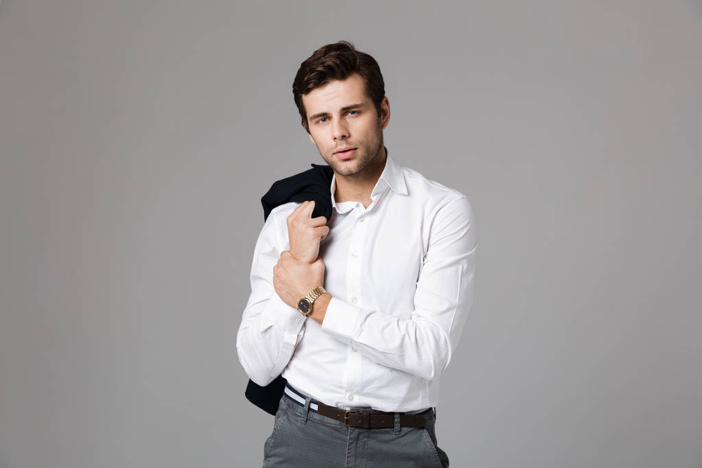Close up portrait of a confident young businessman standing isolated over gray background, looking at camera, holding a jacket - Photo, Image