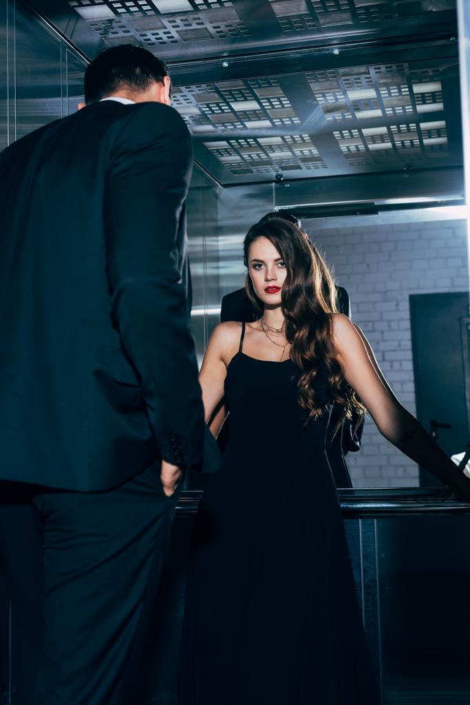 красива спокуслива жінка в чорній сукні дивиться на камеру з чоловіком на передньому плані в ліфті
 - Фото, зображення