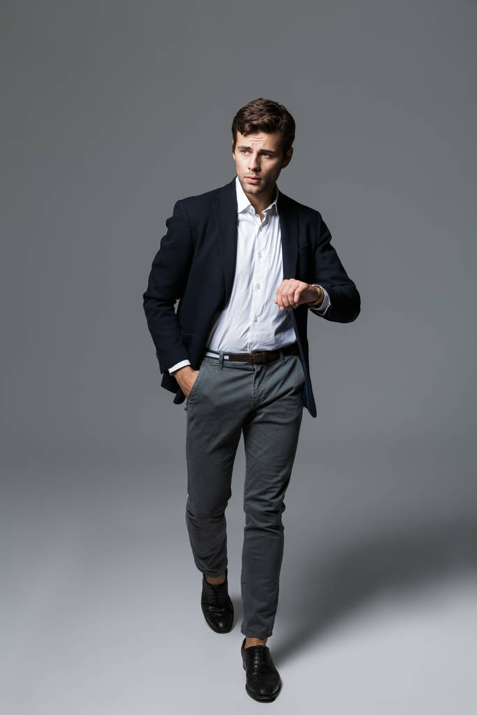 Retrato de larga duración de un joven hombre de negocios guapo vestido con traje aislado sobre fondo gris, posando, comprobando el tiempo, caminando
 - Foto, imagen