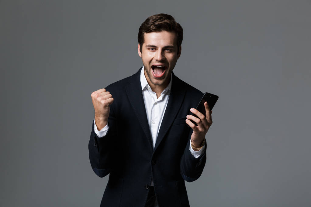 Portret van een vrolijke jonge zakenman gekleed in pak geïsoleerd over grijze achtergrond, houden van mobiele telefoon, vieren - Foto, afbeelding