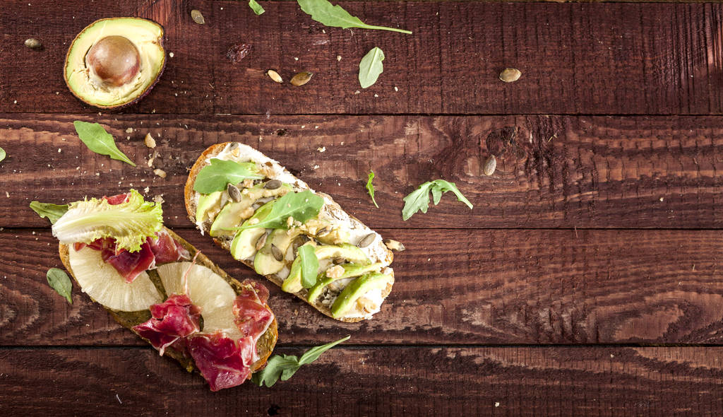 Nahaufnahme von Bruschettas mit Avocado, Mascarpone, Rucolanüssen, Schinken und Ananas auf einem Holztisch. - Foto, Bild