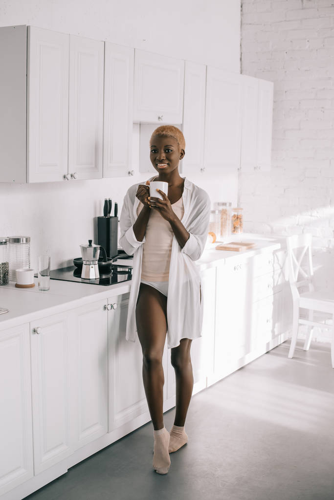 ελκυστική αφρικανική αμερικανική γυναίκα πιτζάμες με φλιτζάνι καφέ στην λευκή κουζίνα  - Φωτογραφία, εικόνα
