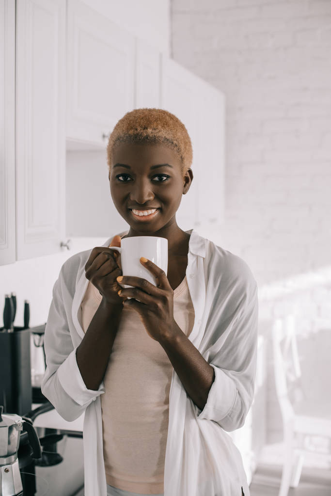χαρούμενα αφρικανική αμερικανική γυναίκα με φλιτζάνι καφέ στην λευκή κουζίνα  - Φωτογραφία, εικόνα