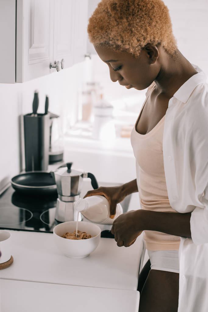 krásná africká americká žena vylévání mléka v misce s kukuřičnými lupínky v kuchyni  - Fotografie, Obrázek