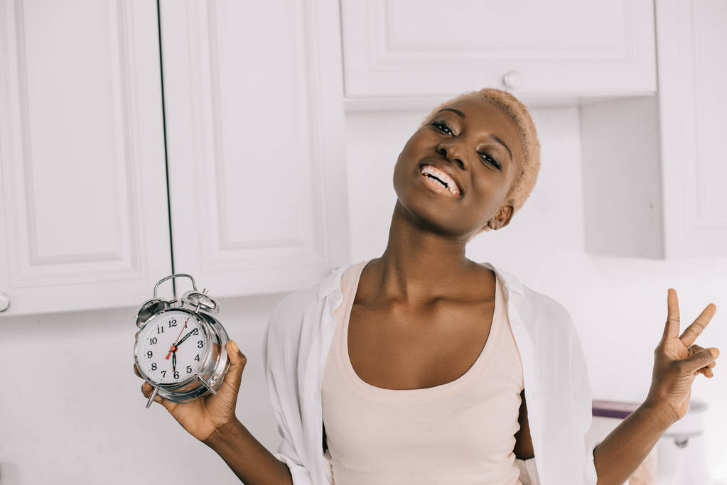 веселая африканская американка держит часы и показывает знак мира
 - Фото, изображение