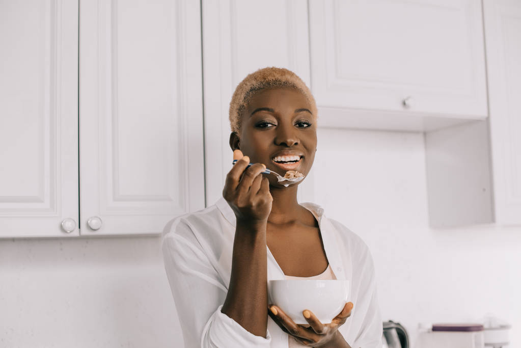 alegre afroamericana mujer comiendo copos de maíz en blanco cocina
  - Foto, imagen