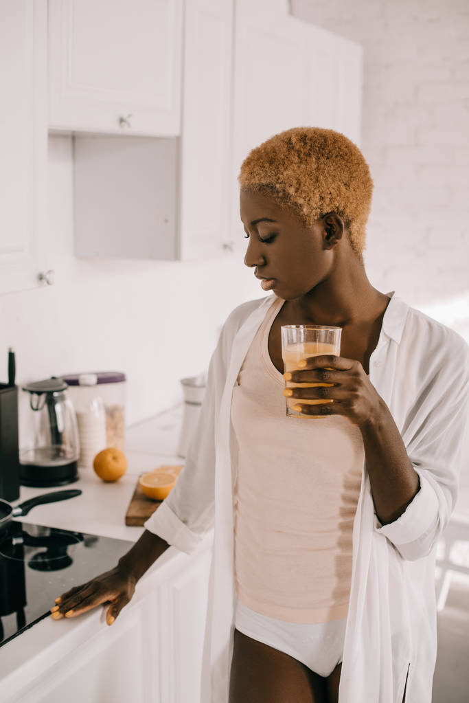 doordachte Afro-Amerikaanse vrouw met glas sinaasappelsap in keuken  - Foto, afbeelding