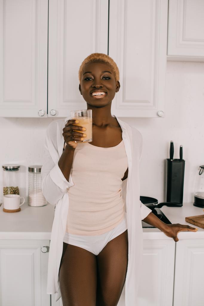 ευτυχισμένος αφρικανική αμερικανική γυναίκα που δείχνει ποτήρι χυμό πορτοκαλιού στο λευκό κουζίνα  - Φωτογραφία, εικόνα