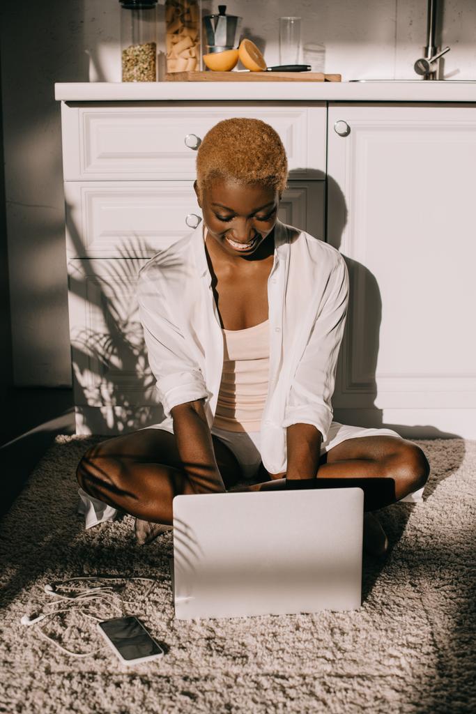  african american woman sitting on carpet and typing on laptop in white kitchen - Photo, Image