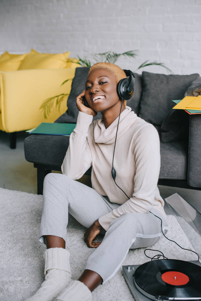 prachtige Afrikaanse Amerikaanse vrouw zittend op een tapijt en luisteren muziek in hoofdtelefoons - Foto, afbeelding