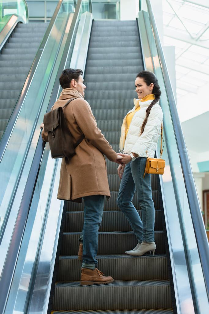 sonriente pareja en ropa de abrigo cogido de la mano, mirándose unos a otros y subiendo en escaleras mecánicas
 - Foto, imagen