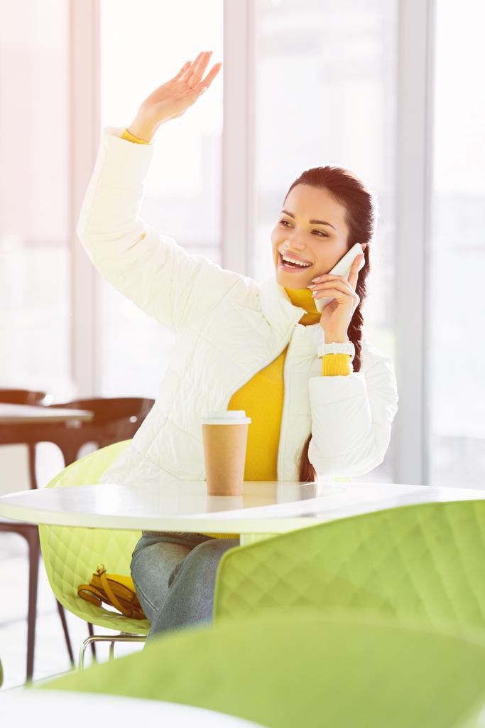 усміхнена дівчина сидить за столом, розмовляє на смартфоні і махає рукою в кафе
 - Фото, зображення