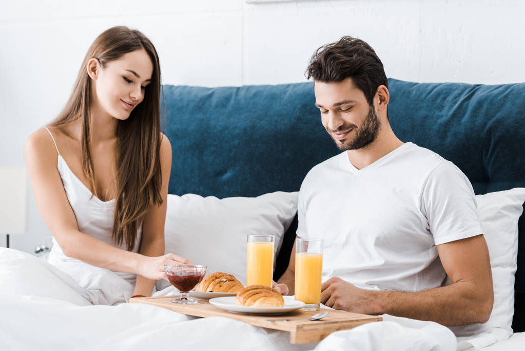 pareja joven sentada en la cama con desayuno en bandeja de madera
 - Foto, imagen