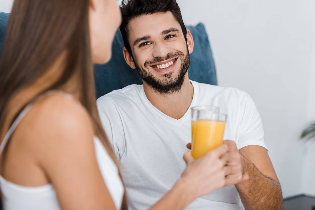 Selektywny fokus przystojny uśmiechnięty mężczyzna ogląda dziewczyna i trzymając szklankę soku pomarańczowego - Zdjęcie, obraz