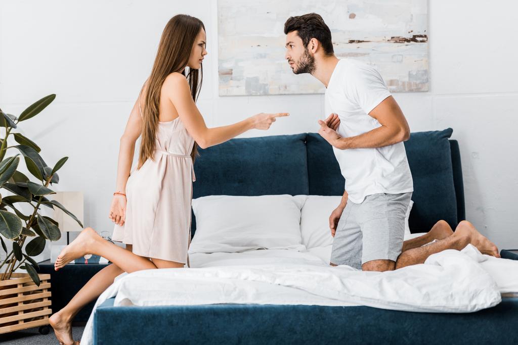 junge attraktive Frau zeigt wütend mit dem Finger auf gestressten gestikulierenden Mann, während er auf dem Bett steht - Foto, Bild