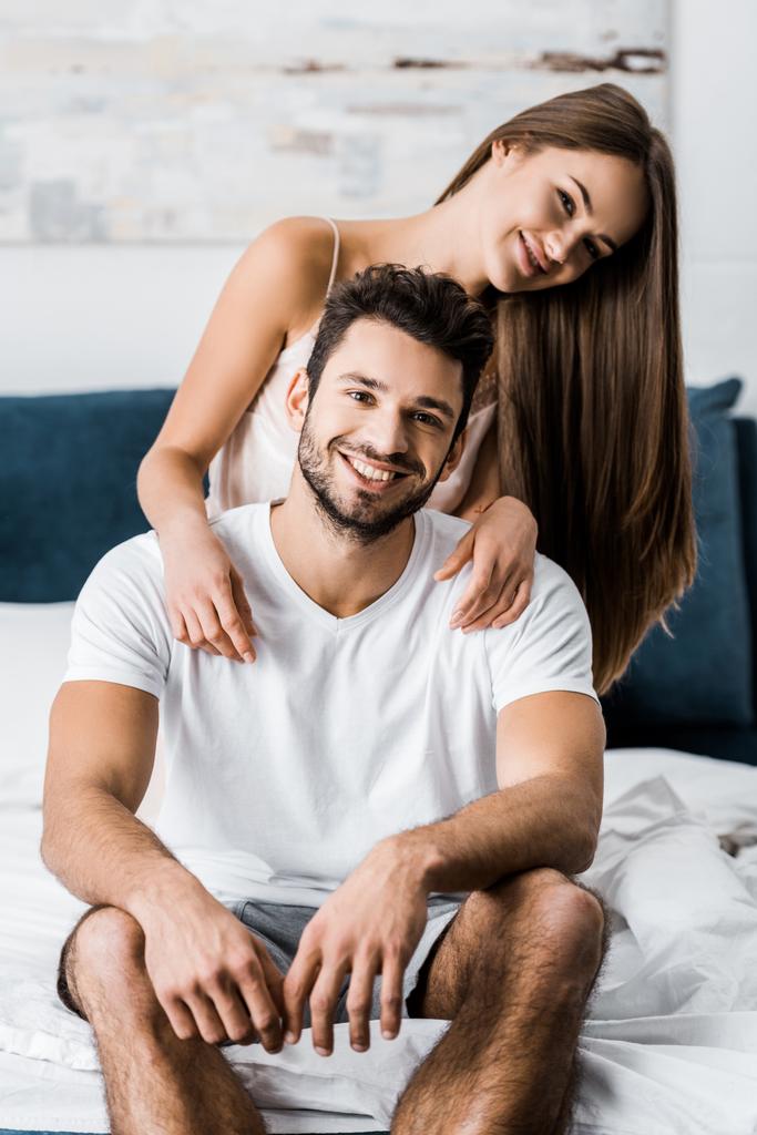 junges attraktives Mädchen umarmt fröhlichen Mann, während es auf dem Bett sitzt - Foto, Bild