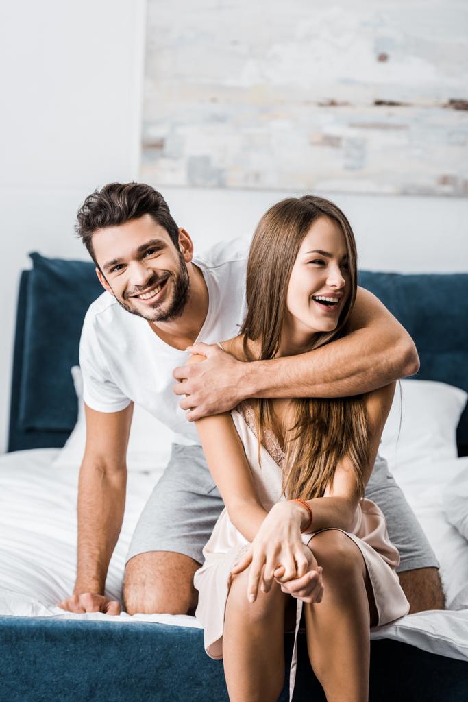 νεαρός αγκαλιάζει γυναίκα με ένα χέρι και χαμογελώντας, ενώ κάθονται στο κρεβάτι - Φωτογραφία, εικόνα