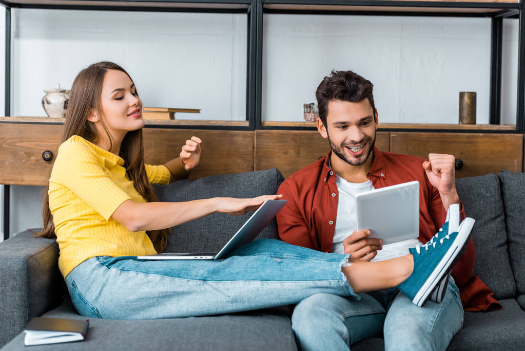 young cheerful couple spending time together on sofa with laptop and digital tablet - Photo, Image