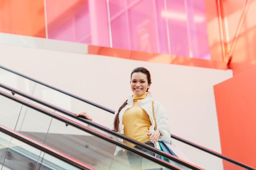 chica atractiva con bolsas de compras sonriendo y mirando a la cámara en la escalera mecánica
 - Foto, Imagen