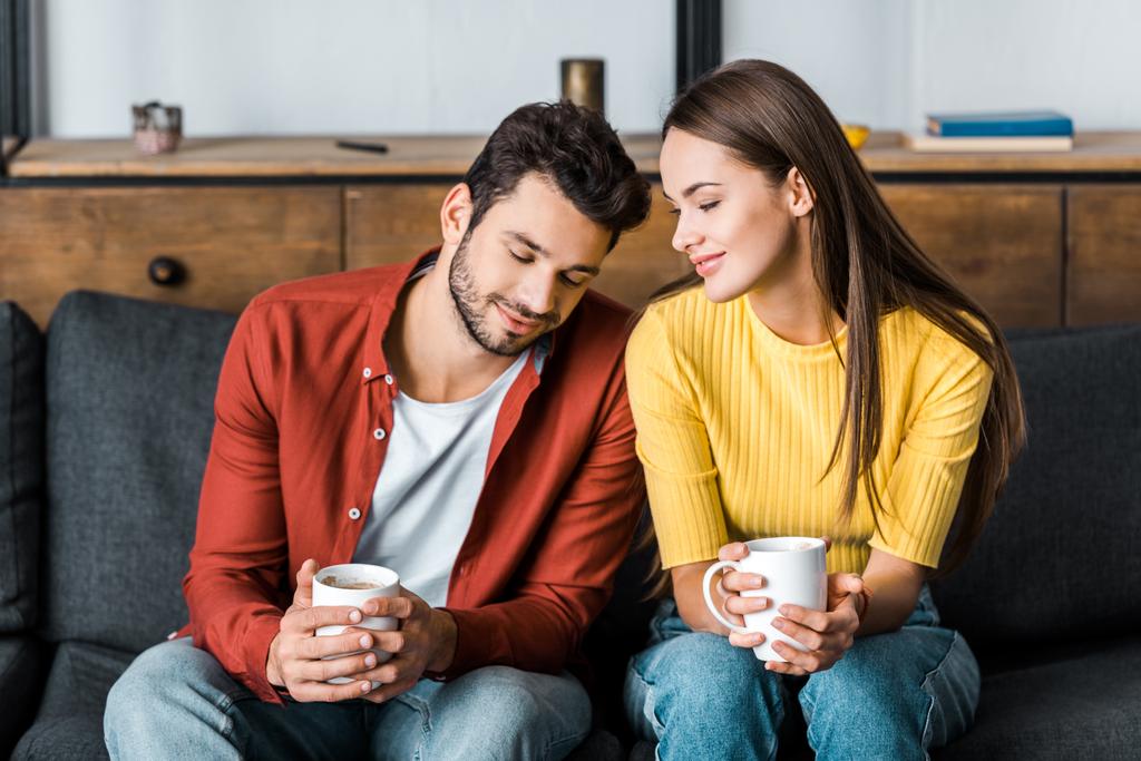 happy couple sitting on sofa and holding cups with drinks - Photo, Image