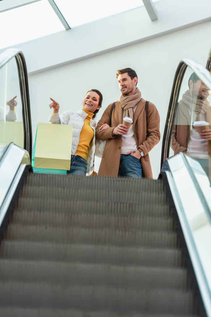 menina atraente com sacos de compras apontando com o dedo e segurando a mão de homem bonito com copo de papel no topo da escada rolante
  - Foto, Imagem