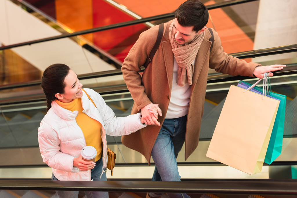 sorrindo homem com sacos de compras e menina bonita com copo descartável olhando uns para os outros na escada rolante
 - Foto, Imagem