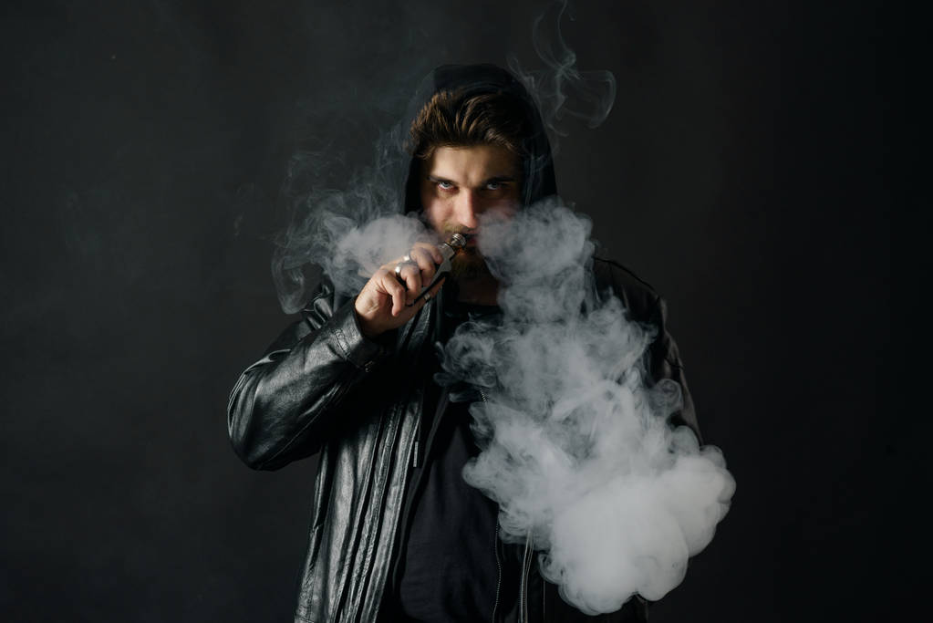 黒のスタジオの背景で蒸気を吐き出す vaping mod を持つ男。男のひげを生やした電子タバコの喫煙をたばこをやめる。蒸気と代替ニコチン無料喫煙コンセプト - 写真・画像