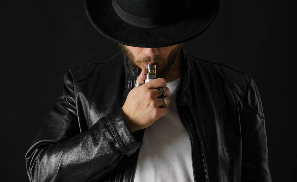 Hombre con vapeo mod exhalando vapor en el fondo del estudio negro. Un tipo barbudo fumando cigarrillo electrónico para dejar el tabaco. Concepto de fumador libre de vapores y nicotina alternativa
 - Foto, Imagen
