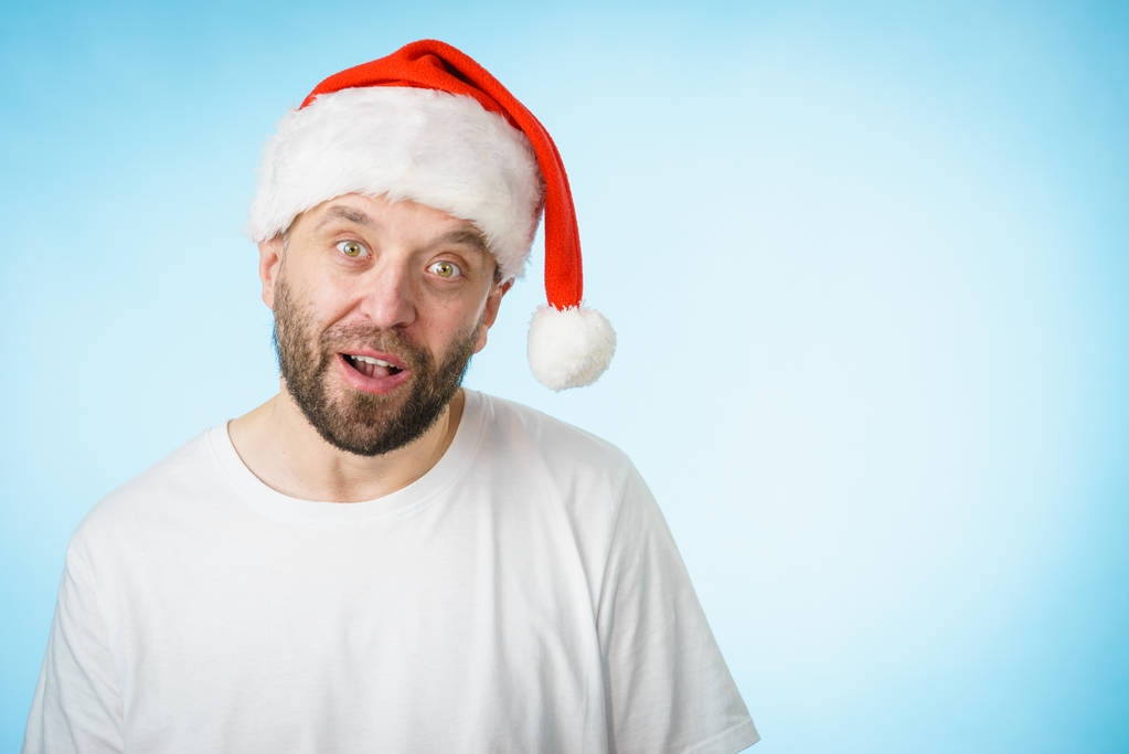 Бородатый мужчина в шляпе Санта Клауса удивлённое выражение лица, вид сбоку на синий с копировальным пространством. Время Рождества
. - Фото, изображение