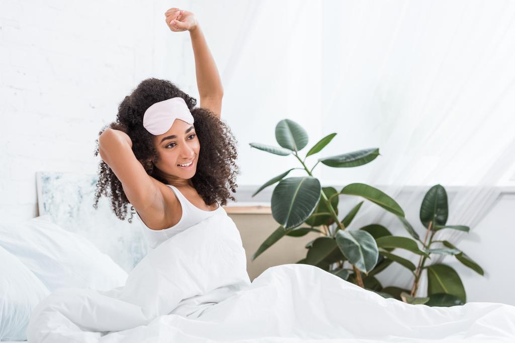 freudige junge afrikanisch-amerikanische Frau mit Augenbinde auf der Stirn, die sich morgens zu Hause im Bett streckt - Foto, Bild