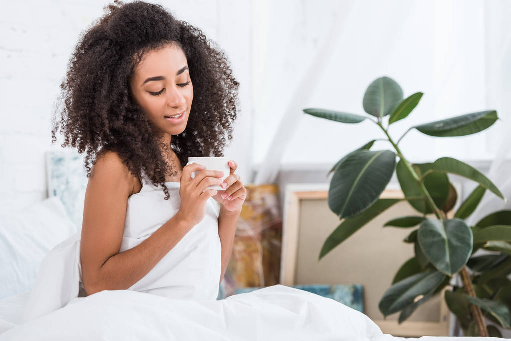 hermosa chica afroamericana rizada sosteniendo taza de café en la cama durante la mañana en casa
 - Foto, Imagen