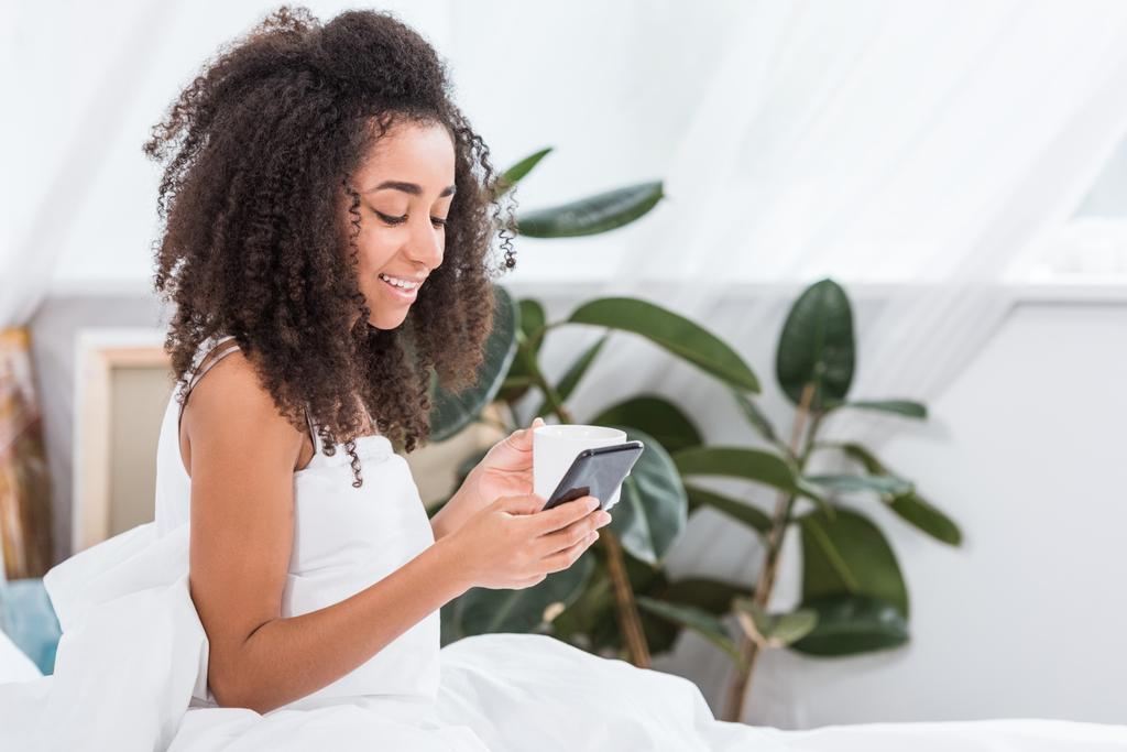 mujer joven afroamericana feliz con taza de café usando teléfono inteligente en la cama durante el horario de la mañana en casa
 - Foto, imagen
