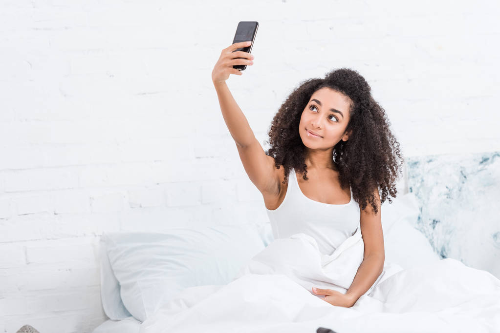 atractiva chica afroamericana tomando selfie en el teléfono inteligente en la cama durante la mañana en casa
 - Foto, imagen