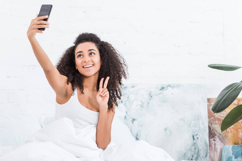 lächelnde Afroamerikanerin zeigt Friedenszeichen und macht morgens zu Hause ein Selfie mit dem Smartphone im Bett - Foto, Bild