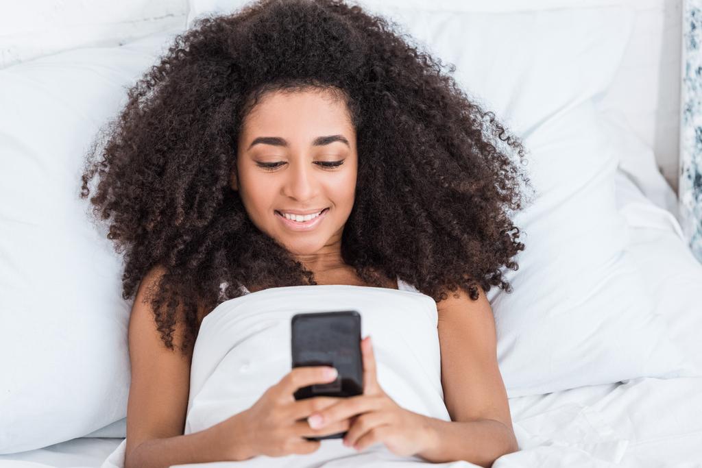 szczęśliwy african american kobieta młody smartfona w łóżku w domu w czasie rano - Zdjęcie, obraz