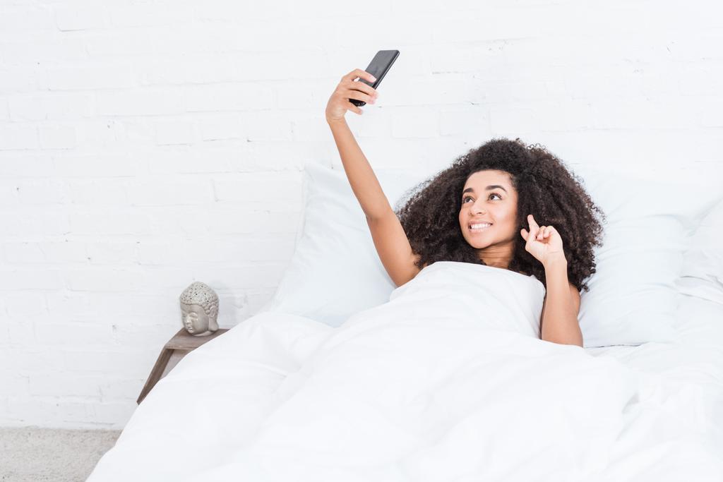 hermosa chica afroamericana mostrando señal de paz y tomando selfie en el teléfono inteligente en la cama durante la mañana en casa
 - Foto, imagen