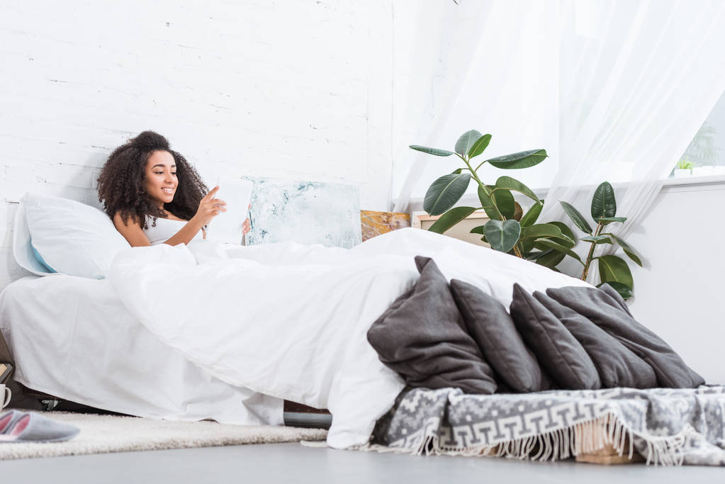 Selektywny fokus z african american kobieta młody za pomocą cyfrowego tabletu w łóżku w domu w czasie rano - Zdjęcie, obraz
