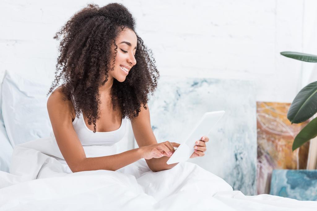 щаслива афроамериканська молода жінка, використовуючи цифровий планшет в ліжку в ранковий час вдома
 - Фото, зображення